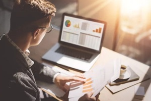 finance market analyst in eyeglasses working at sunny office on laptop while sitting at wooden table